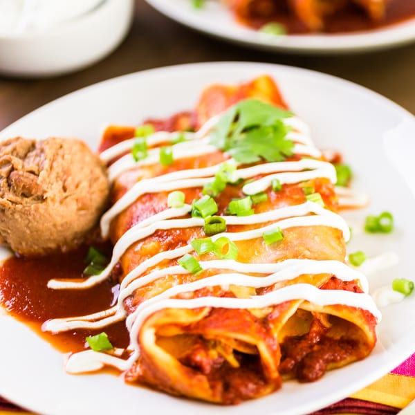 Three enchiladas with red sauce, sour cream, green onions, and cilantro, served with refried beans.