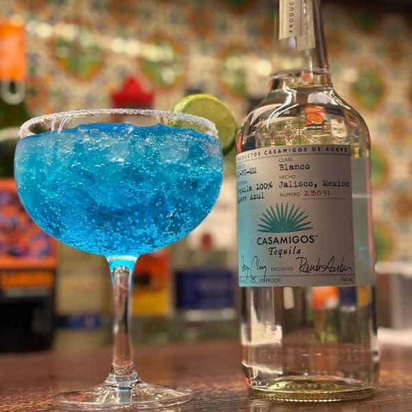 A vibrant blue margarita in a stemmed glass with a lime wedge and a bottle of Casamigos Tequila.